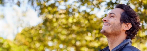 A man standing outside in nature feeling content and happy, smiling and looking to the sky.